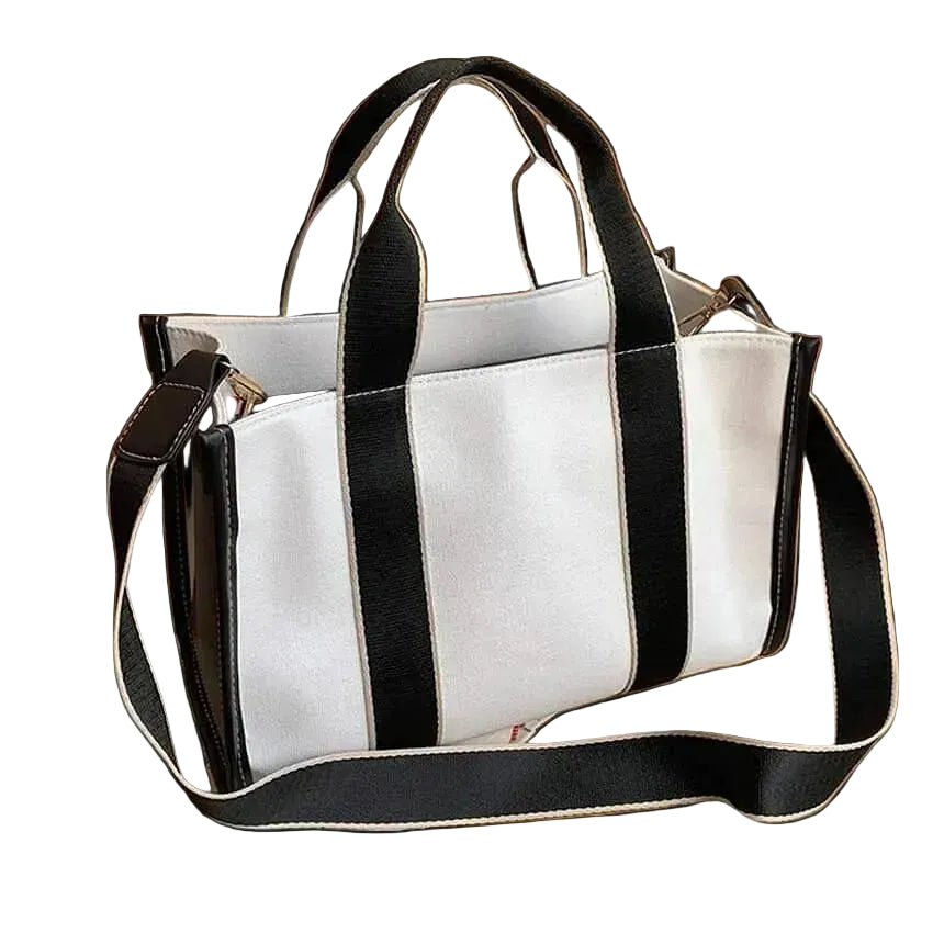 A white canvas tote bag with bold black vertical stripes, featuring dual top handles and a matching adjustable shoulder strap, styled on a wooden tabletop with a book and a white vase.