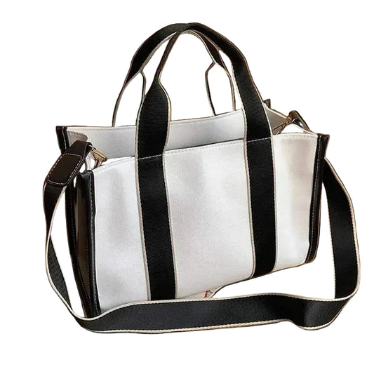 A white canvas tote bag with bold black vertical stripes, featuring dual top handles and a matching adjustable shoulder strap, styled on a wooden tabletop with a book and a white vase.